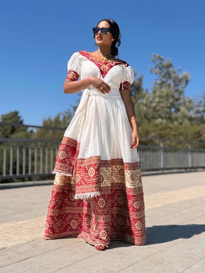 Woman wearing a white and red patterned Ethiopian cultural dress standing outdoors on a sunny day.