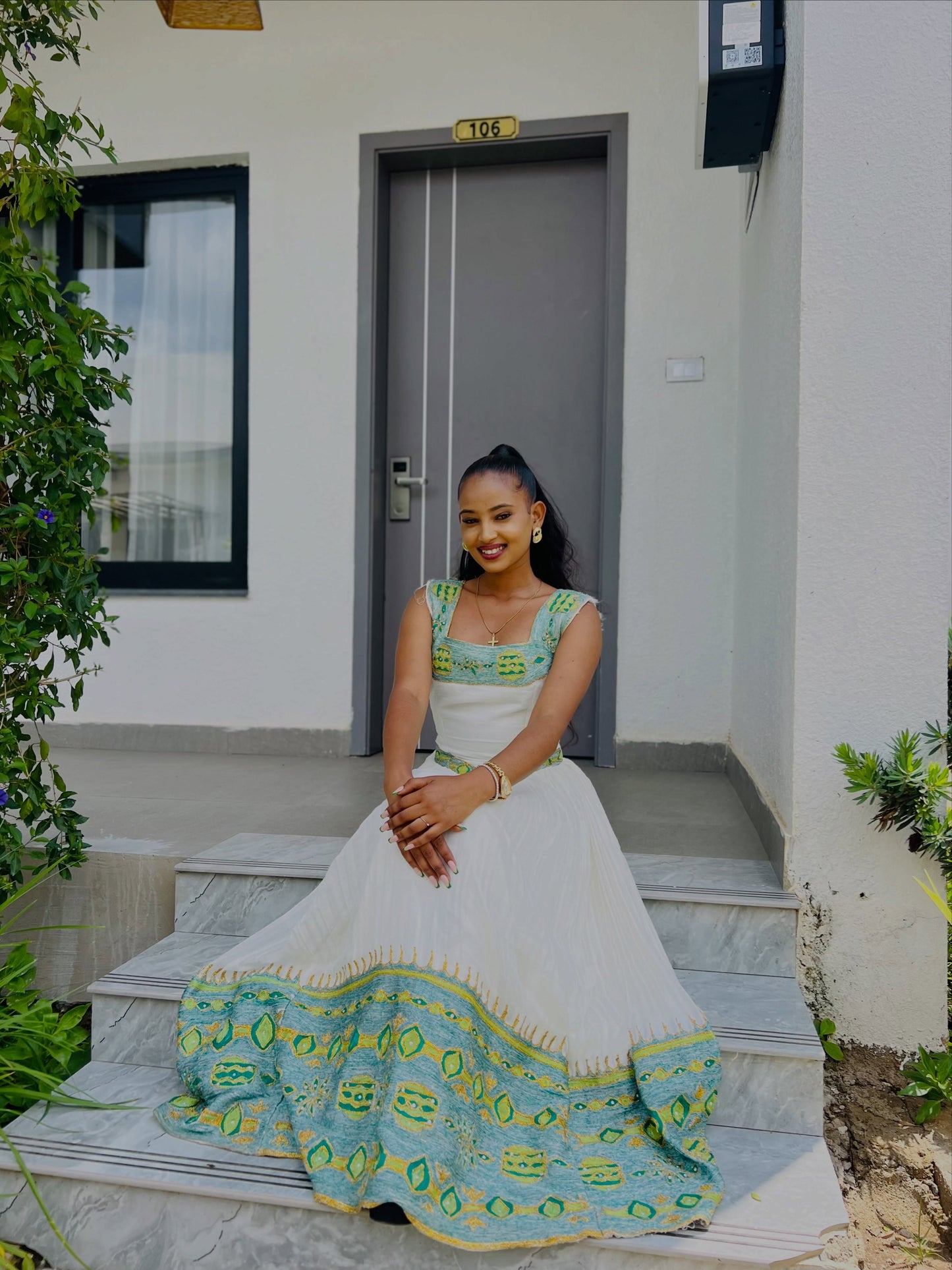 A woman sits on outdoor stone steps in front of a modern building, wearing a sleeveless Ethiopian Kemis with a turquoise and green patterned bodice and hem.