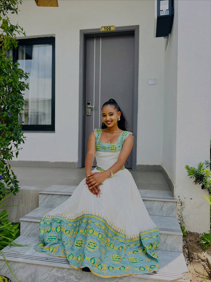 A woman sits on outdoor stone steps in front of a modern building, wearing a sleeveless Ethiopian Kemis with a turquoise and green patterned bodice and hem.