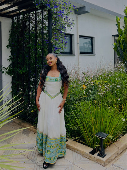 A woman wearing a white Ethiopian Kemis with green and turquoise patterns, standing in a garden courtyard.