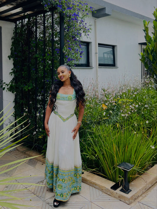 A woman wearing a white Ethiopian Kemis with green and turquoise patterns, standing in a garden courtyard.