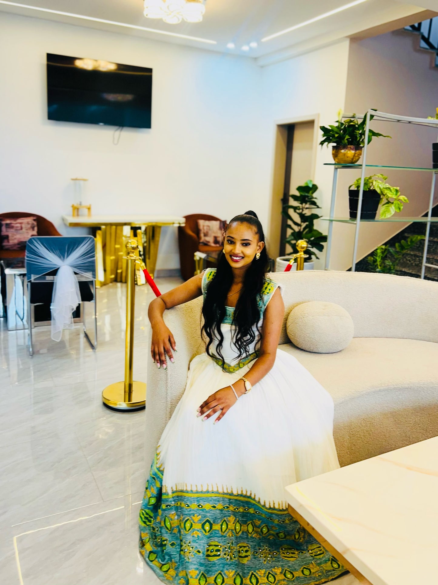 A woman wearing a Ethiopian Kemis with green and gold patterns sits on a beige sofa in a modern, upscale lounge setting.