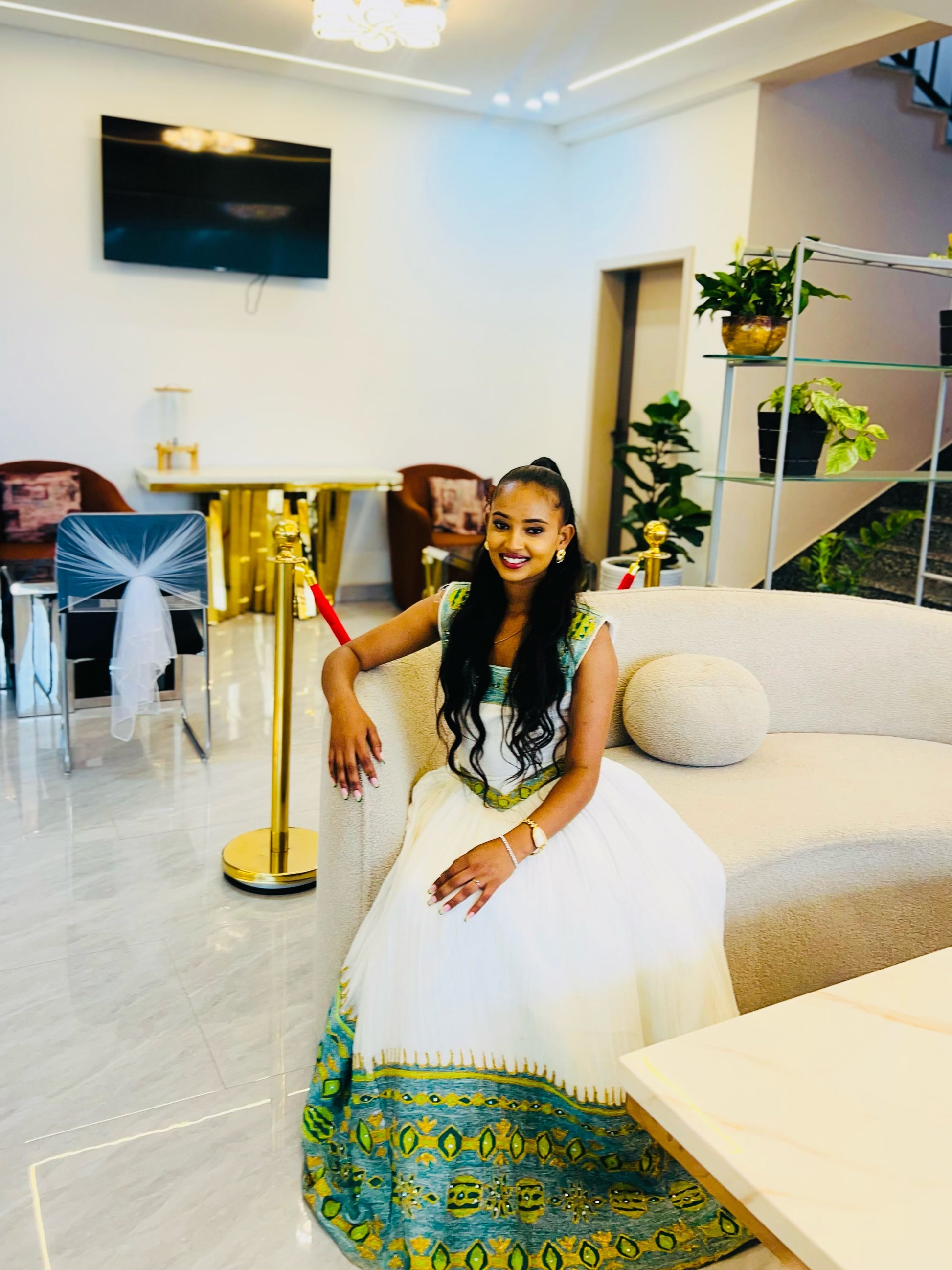 A woman wearing a Ethiopian Kemis with green and gold patterns sits on a beige sofa in a modern, upscale lounge setting.