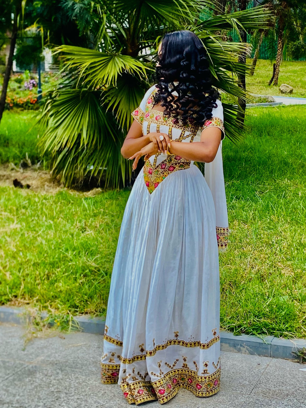 A woman wearing a white traditional Habesha Store with colorful floral embroidery, photographed from behind in an outdoor park setting with green grass and palm trees.