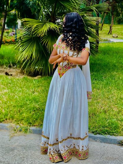 A woman wearing a white traditional Habesha Store with colorful floral embroidery, photographed from behind in an outdoor park setting with green grass and palm trees.