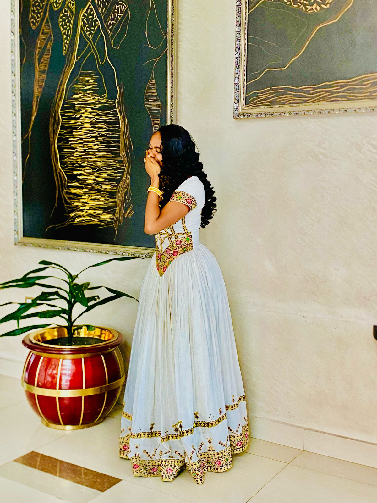 A woman wearing a white traditional Habesha Store with gold and red embroidery stands in a decorated indoor setting with paintings and a potted plant.