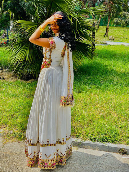 A woman wearing a white traditional Habesha Store with colorful embroidered borders stands outdoors on a paved path with green grass and palm trees in the background.