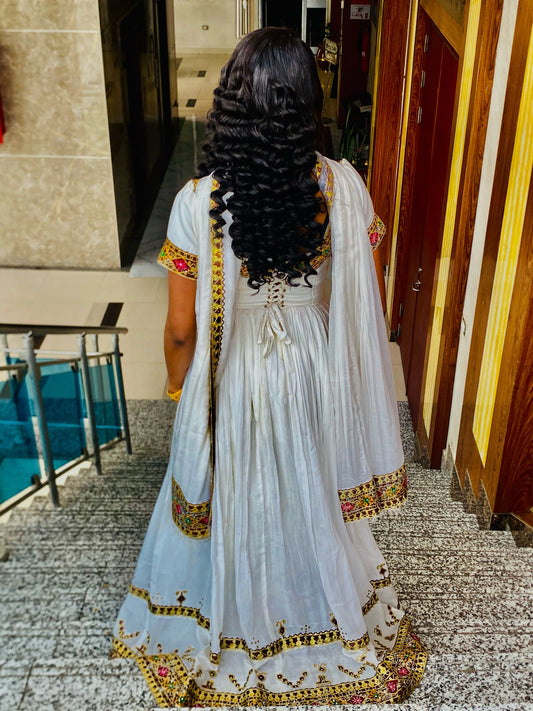 A woman viewed from behind wearing a white traditional Habesha Store with gold and pink embroidered borders and a matching sheer dupatta, standing indoors on a tiled floor.