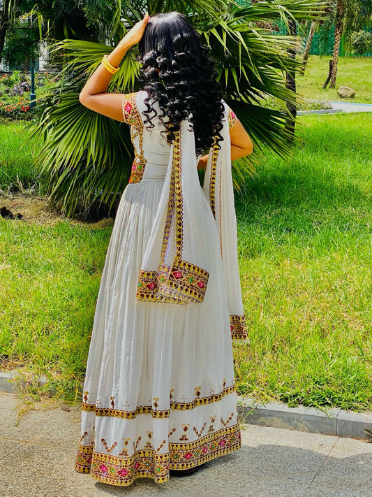 A woman wearing a white traditional ethnic Habesha Store with gold and red embroidery, photographed from behind in an outdoor park setting with green grass and palm leaves.