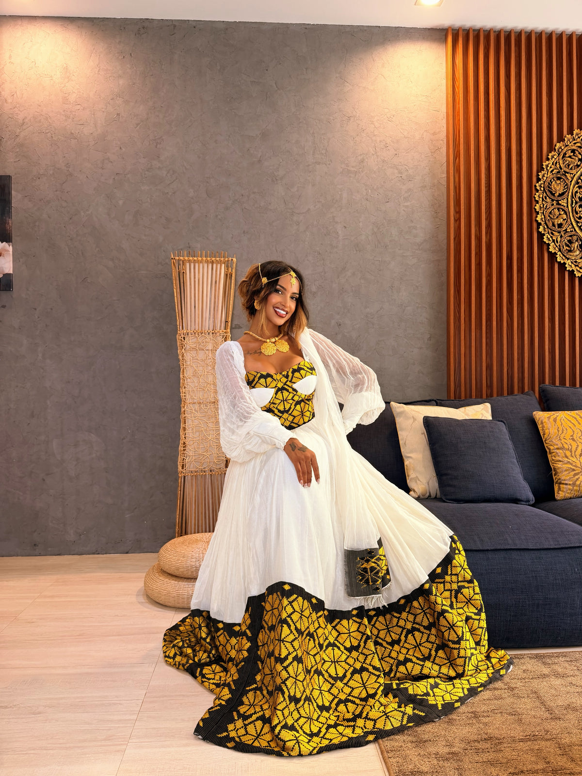 A woman models a traditional African Ethiopia clothes with a white tulle overlay and a yellow, black, and green patterned waist and skirt, posed in a modern living room setting.