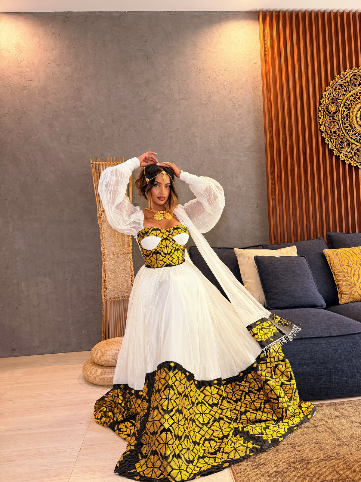 A woman models a traditional South Asian Ethiopia clothes featuring a white tulle skirt with a bold yellow and black geometric patterned hem, paired with a matching yellow and black embroidered bodice and sheer white balloon sleeves, in a modern living room setting.
