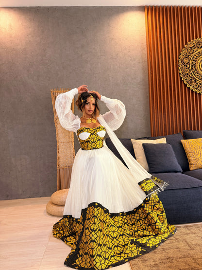 A woman models a traditional South Asian Ethiopia clothes featuring a white tulle skirt with a bold yellow and black geometric patterned hem, paired with a matching yellow and black embroidered bodice and sheer white balloon sleeves, in a modern living room setting.