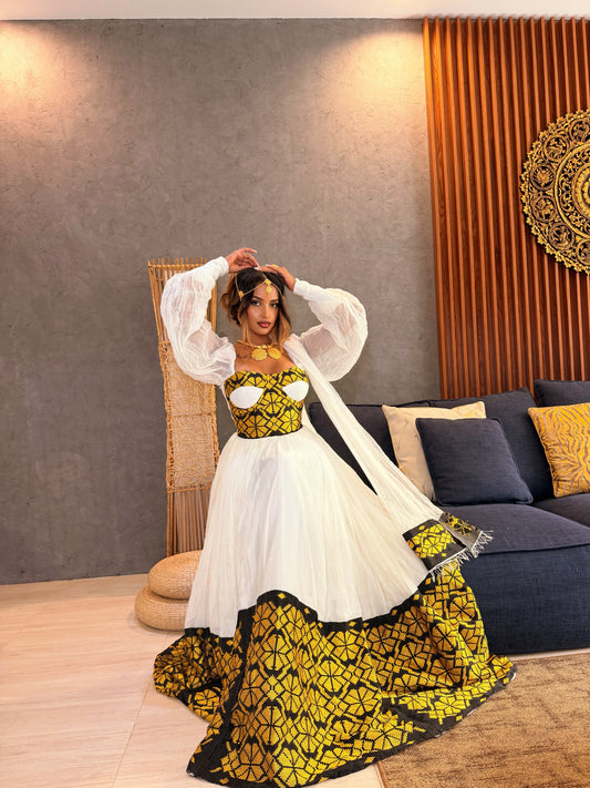 A woman models a traditional South Asian Ethiopia clothes featuring a white tulle skirt with a bold yellow and black geometric patterned hem, paired with a matching yellow and black embroidered bodice and sheer white balloon sleeves, in a modern living room setting.