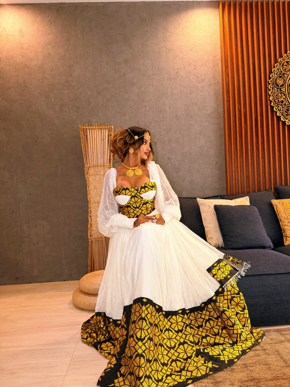 A woman wearing a traditional African Ethiopia clothes with a white tulle skirt and a yellow and black patterned bodice and hem, seated in a modern living room setting.