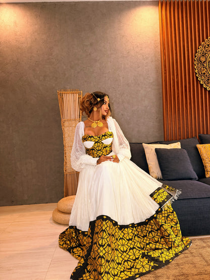 A woman wearing a traditional African Ethiopia clothes with a white tulle skirt and a yellow and black patterned bodice and hem, seated in a modern living room setting.