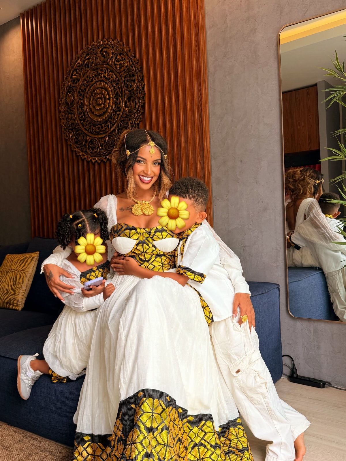 A woman and two children wearing matching white and yellow traditional African Ethiopia clothes pose together on a blue sofa in a modern living room. The children's faces are obscured by yellow flower graphics.
