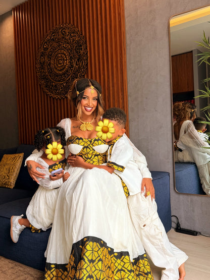 A woman and two children wearing matching white and yellow traditional African Ethiopia clothes pose together on a blue sofa in a modern living room. The children's faces are obscured by yellow flower graphics.