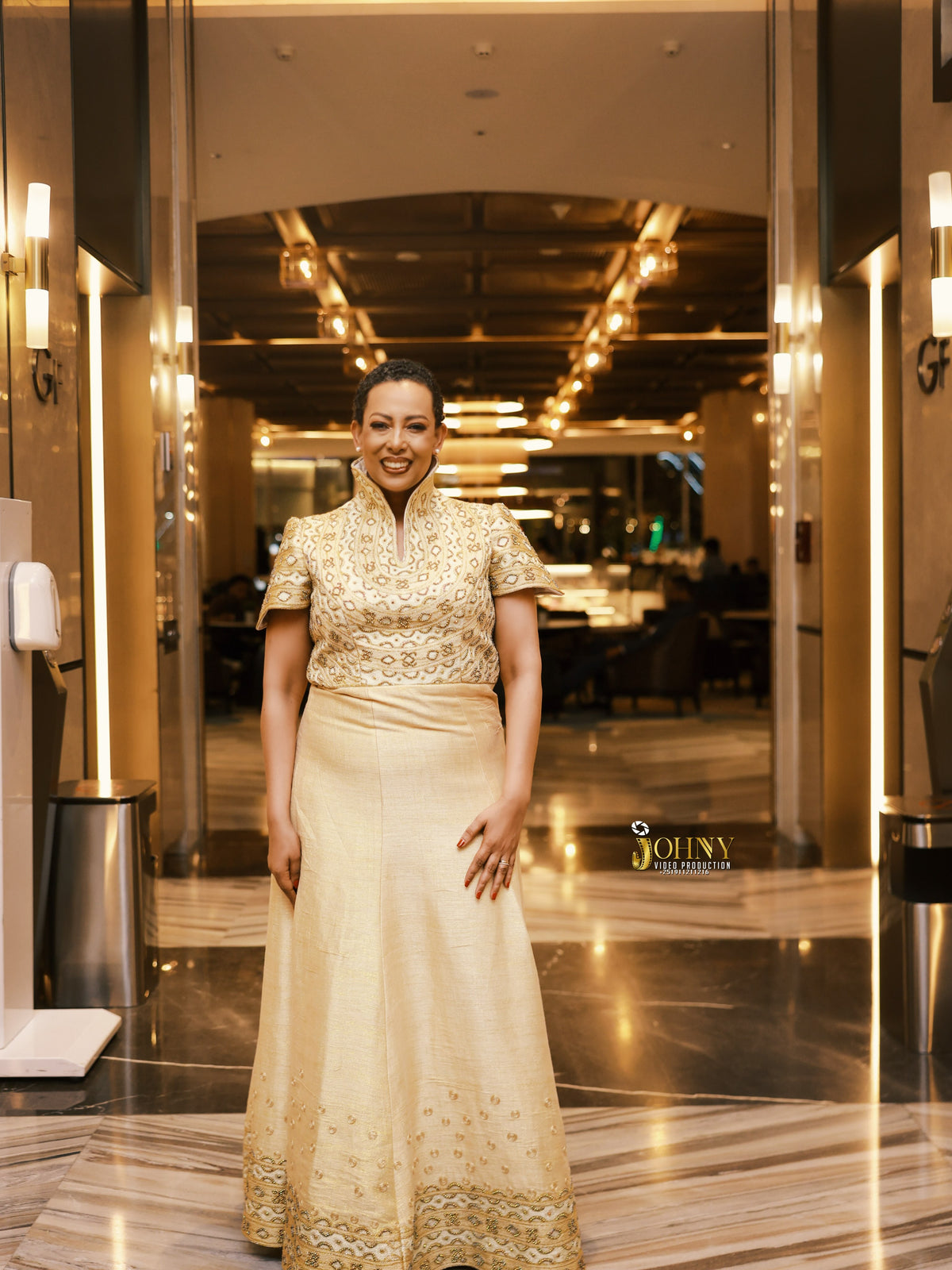 A woman wearing a traditional South Asian formal ethiopian dress for women in beige and gold stands in a hotel lobby. The dress features a heavily embroidered gold bodice with short sleeves and a solid beige skirt.