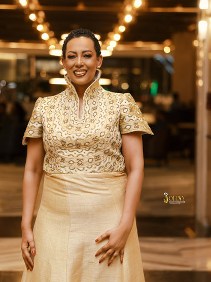 A woman wearing a traditional-style cream and gold embroidered Habesha Kemis  with a high collar and short sleeves, standing in a warmly lit indoor setting.