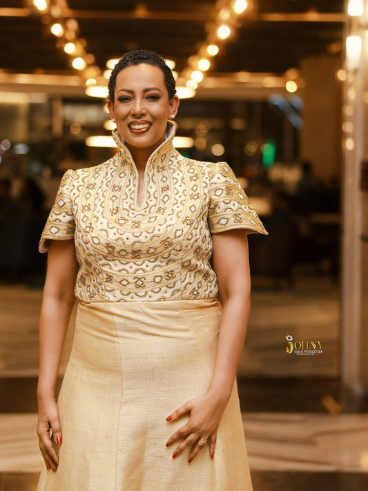 A woman wearing a traditional-style cream and gold embroidered Habesha Kemis  with a high collar and short sleeves, standing in a warmly lit indoor setting.