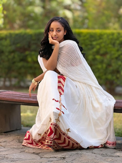Woman in a white Ethiopian cultural dress   with red and gold patterns sitting on a bench outdoors.