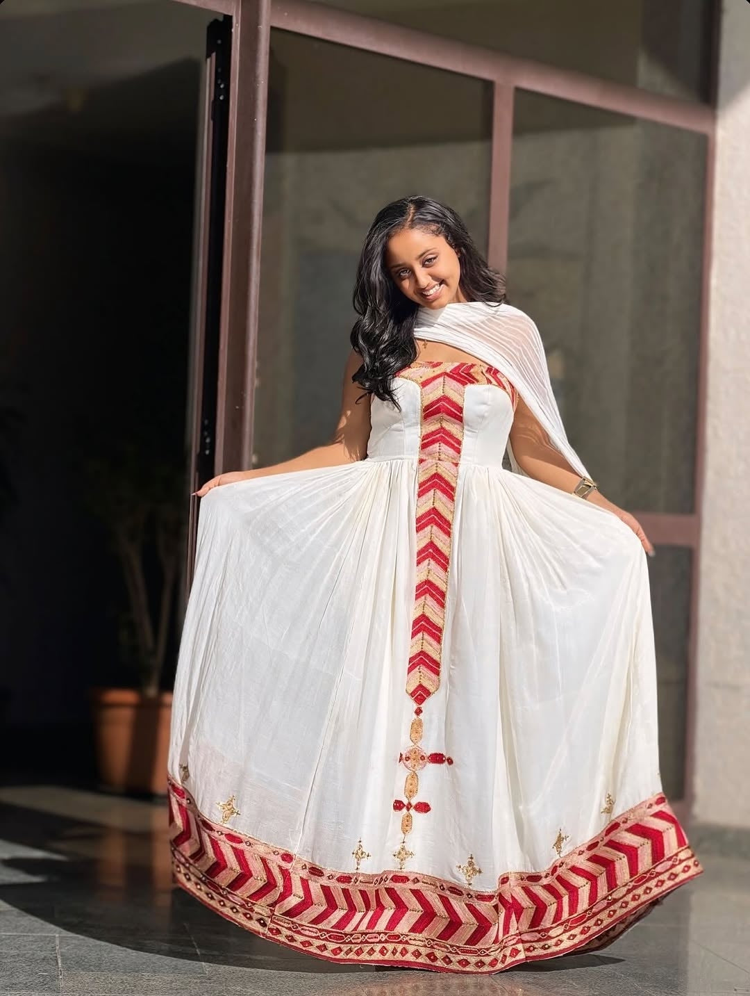 Woman wearing a white Ethiopian cultural dress with red and gold patterns, sitting indoors.
