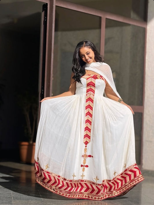 Woman wearing a white Ethiopian cultural dress with red and gold patterns, sitting indoors.