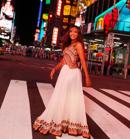 A woman wearing a sleeveless white maxi Habesha Dress with a patterned bodice and a decorative hemline stands on a crosswalk in front of a brightly lit city street at night.