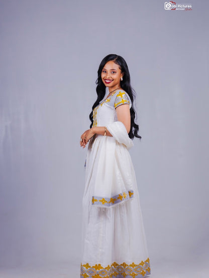 A woman modeling a white traditional habesha kemis with yellow and silver embroidery, standing against a plain light gray studio background.