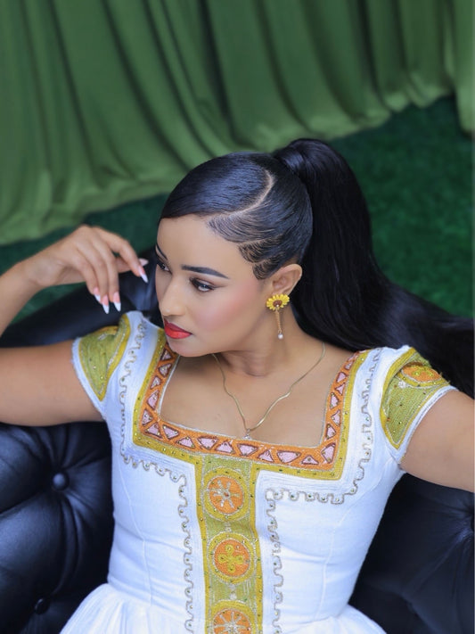 A woman with long black hair in a high ponytail is seated on a black leather sofa, wearing a white Ethiopian shop near me  with ornate gold and green embroidery. She is wearing gold drop earrings and a delicate gold necklace.