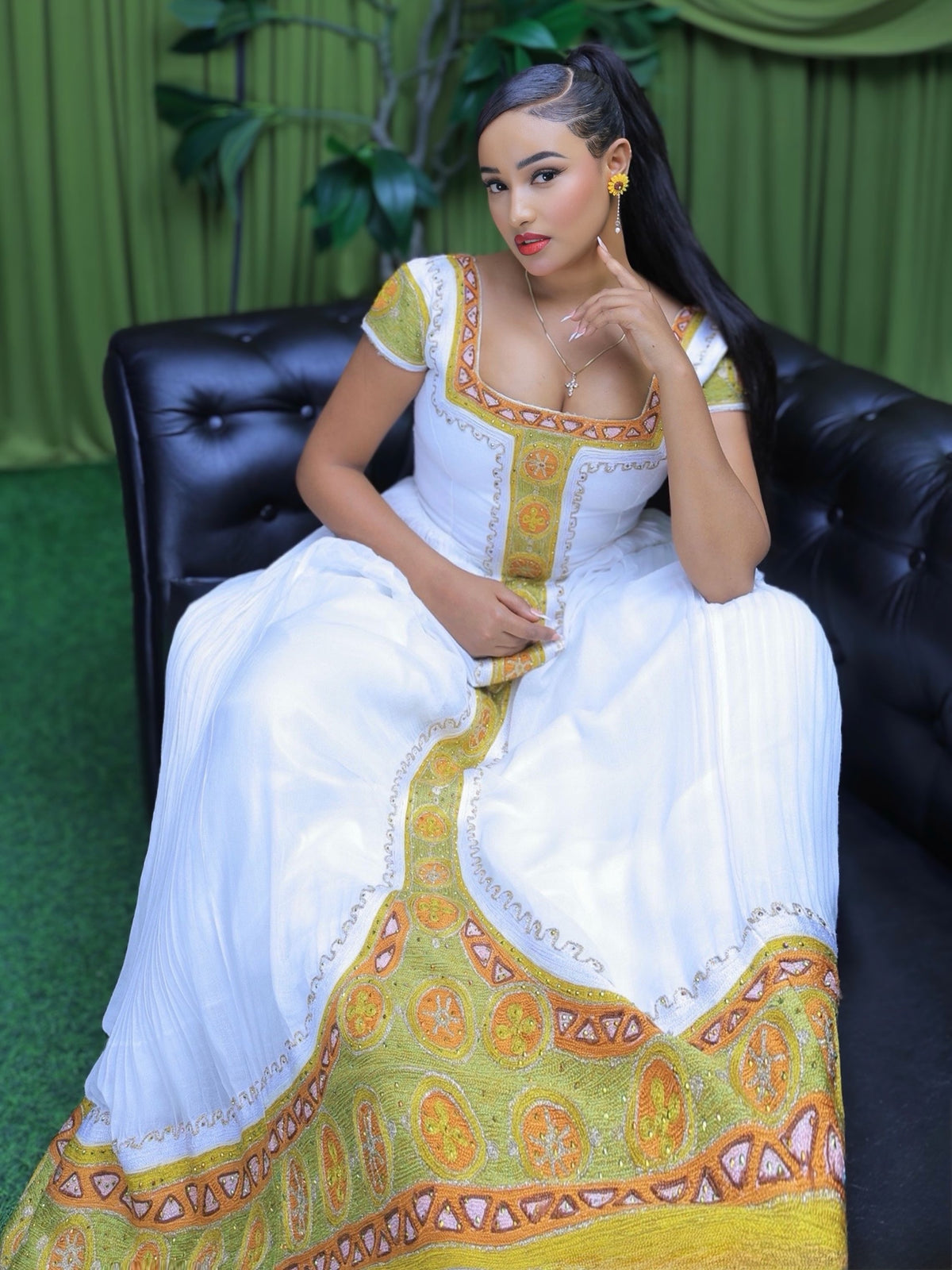 A woman wearing a white traditional Ethiopian shop near me  with ornate yellow and orange embroidery sits on a black leather couch in front of a green curtain backdrop.