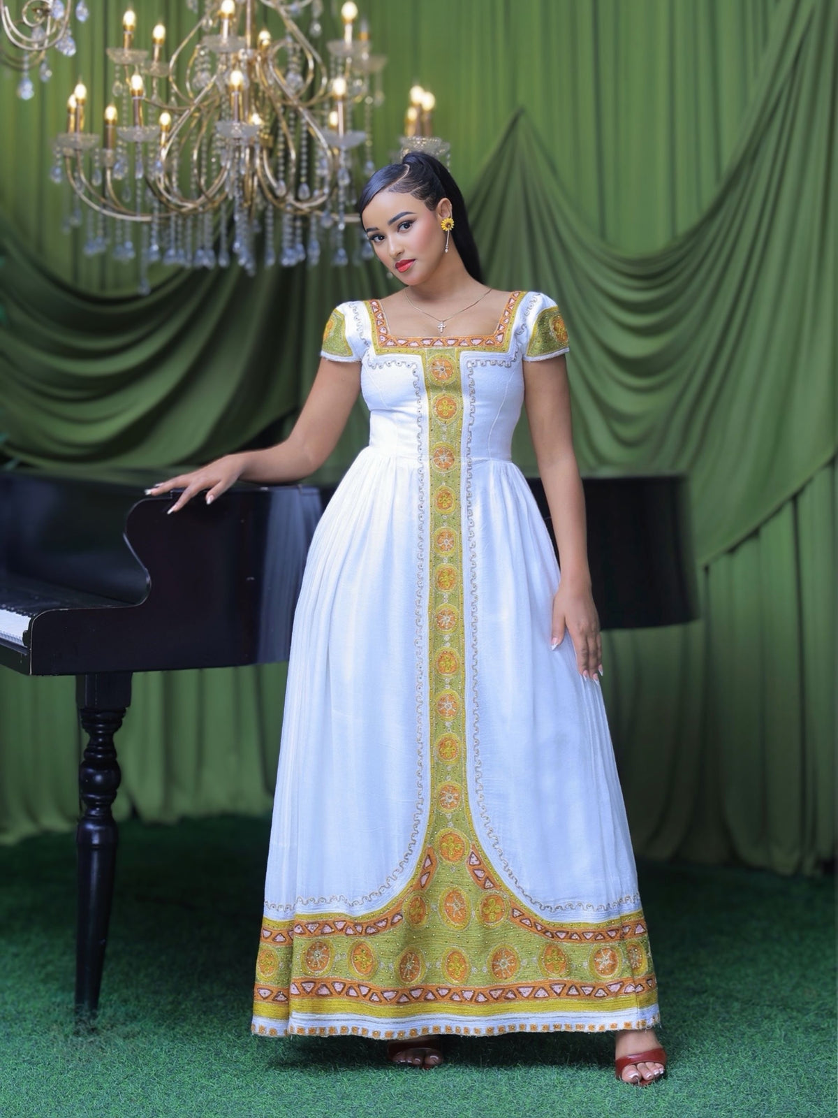 A woman models a white Ethiopian shop near me  with gold and orange traditional embroidery, standing next to a black grand piano against a green draped backdrop.