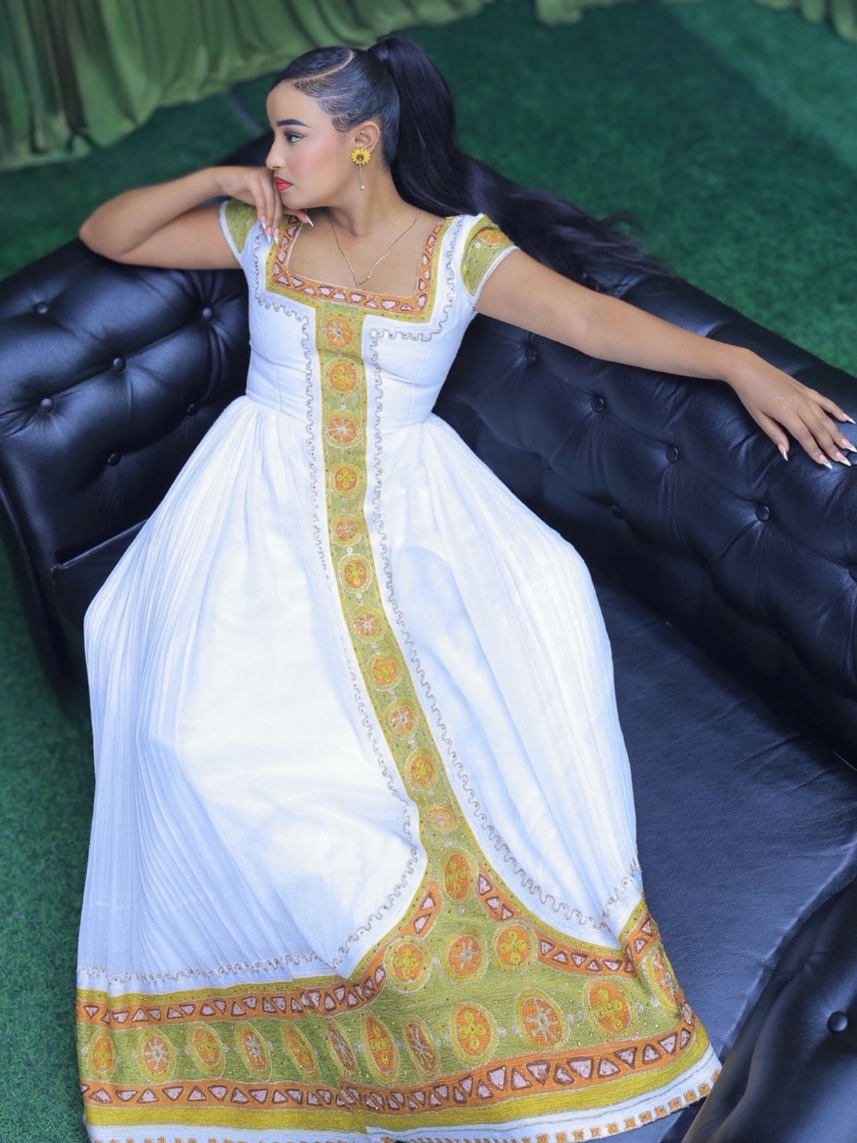 A woman reclining on a black leather sofa wearing a white traditional Ethiopian shop near me  with ornate gold and orange border patterns.