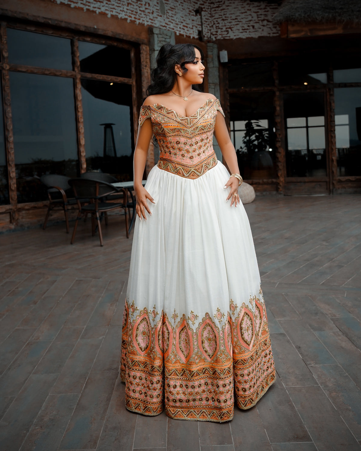 A woman models a traditional Ethiopian Clothing with a white skirt and an orange-gold embroidered bodice and hem, standing on a stone patio.
