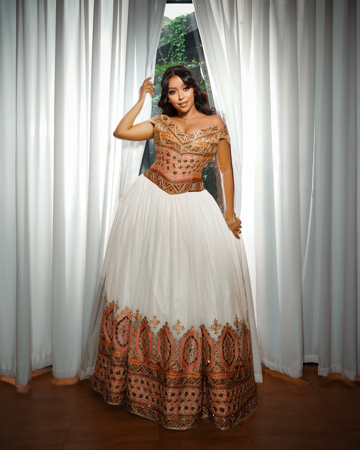 A woman models a traditional Ethiopian Clothing with a white tulle skirt and a bronze-gold embroidered bodice, standing in front of white curtains.