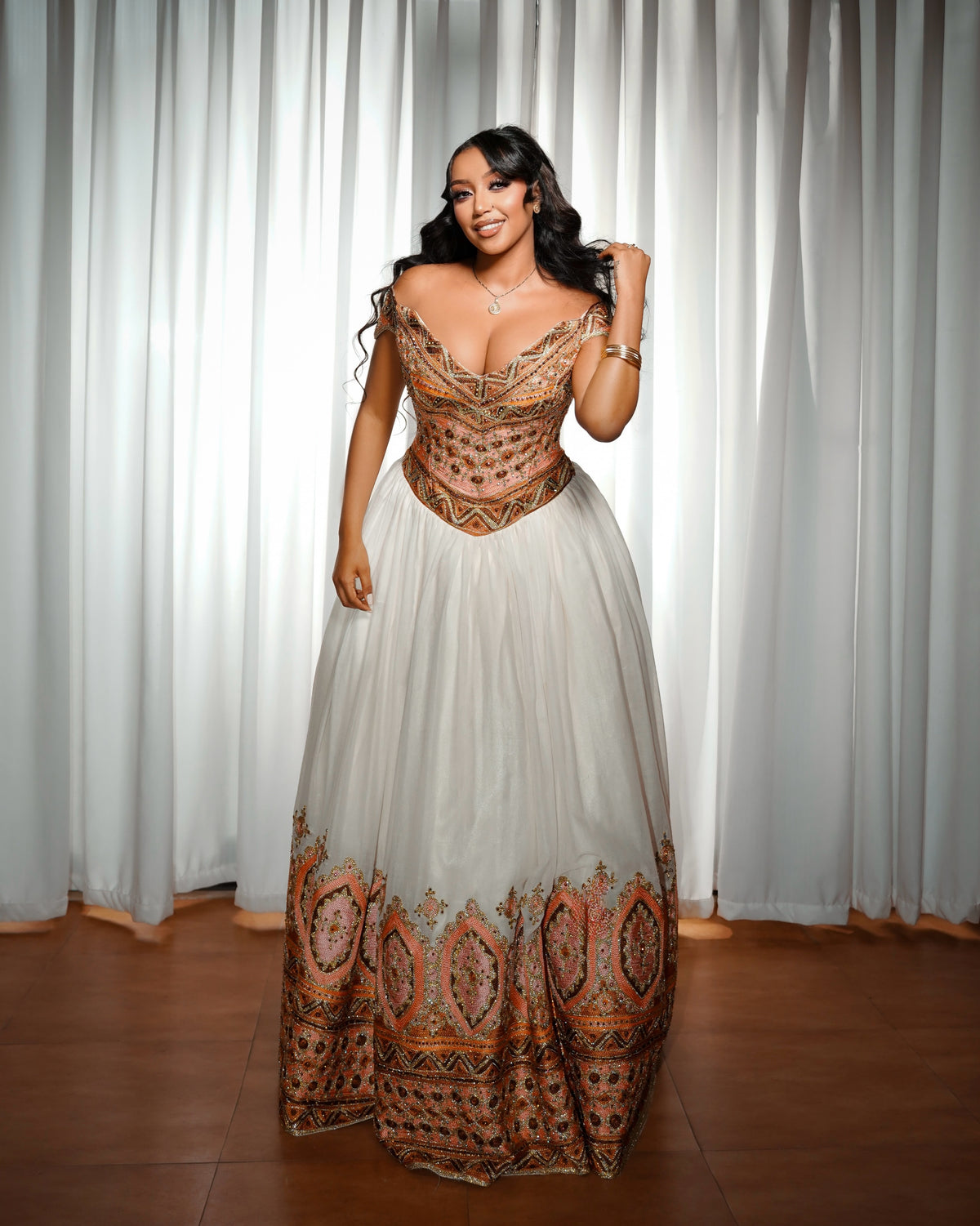 A woman models a floor-length A-line Ethiopian Clothing with a sheer cream tulle skirt and a heavily embellished bronze and gold beaded corset bodice, set against a backdrop of white curtains.