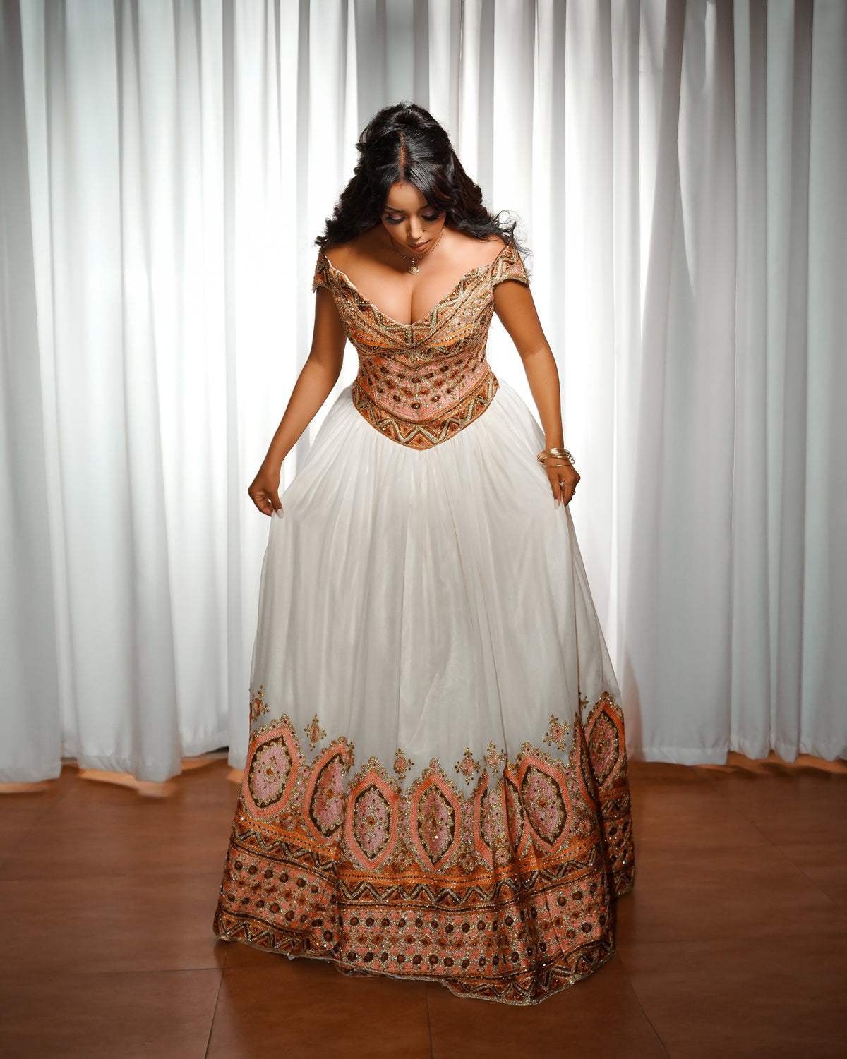 A woman wearing a traditional off-the-shoulder white Ethiopian Clothing with intricate orange, brown, and black embroidered patterns on the bodice and hem, standing in front of white curtains.