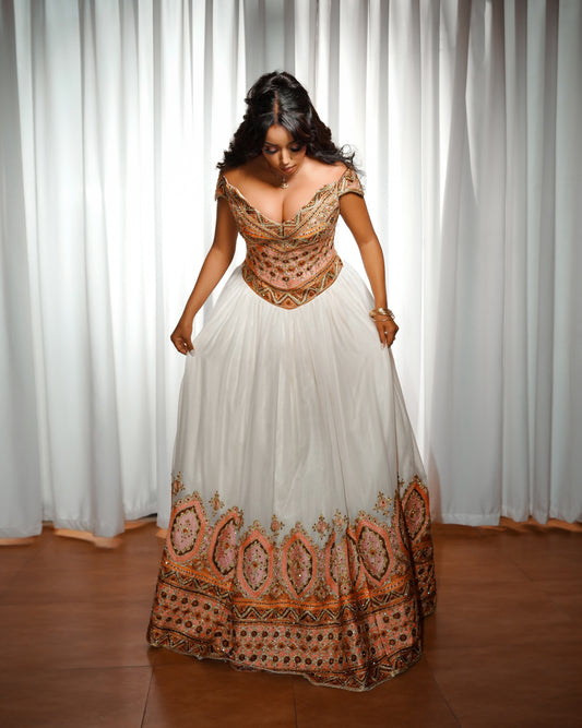 A woman wearing a traditional off-the-shoulder white Ethiopian Clothing with intricate orange, brown, and black embroidered patterns on the bodice and hem, standing in front of white curtains.