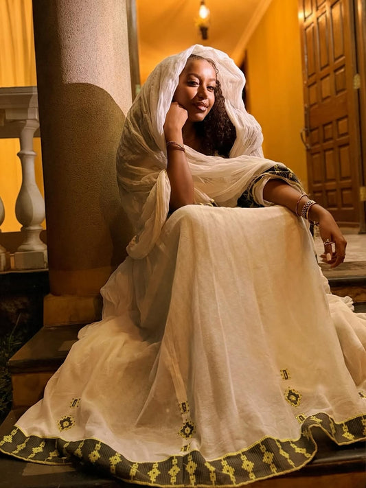 Woman in a white Ethiopian Traditional Dress sitting on steps indoors.