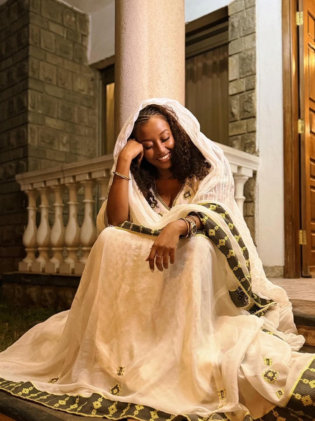 Woman in a white traditional outfit with gold accents sitting on steps.