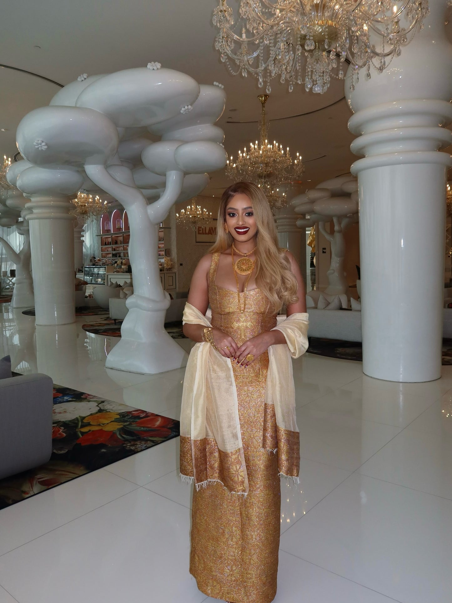 A woman wearing a shimmering gold and beige traditional Habesha kemis with a matching sheer dupatta stands in a luxurious white interior with large columns, chandeliers, and sculptural white trees.