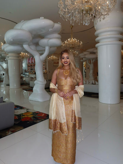 A woman wearing a shimmering gold and beige traditional Habesha kemis with a matching sheer dupatta stands in a luxurious white interior with large columns, chandeliers, and sculptural white trees.