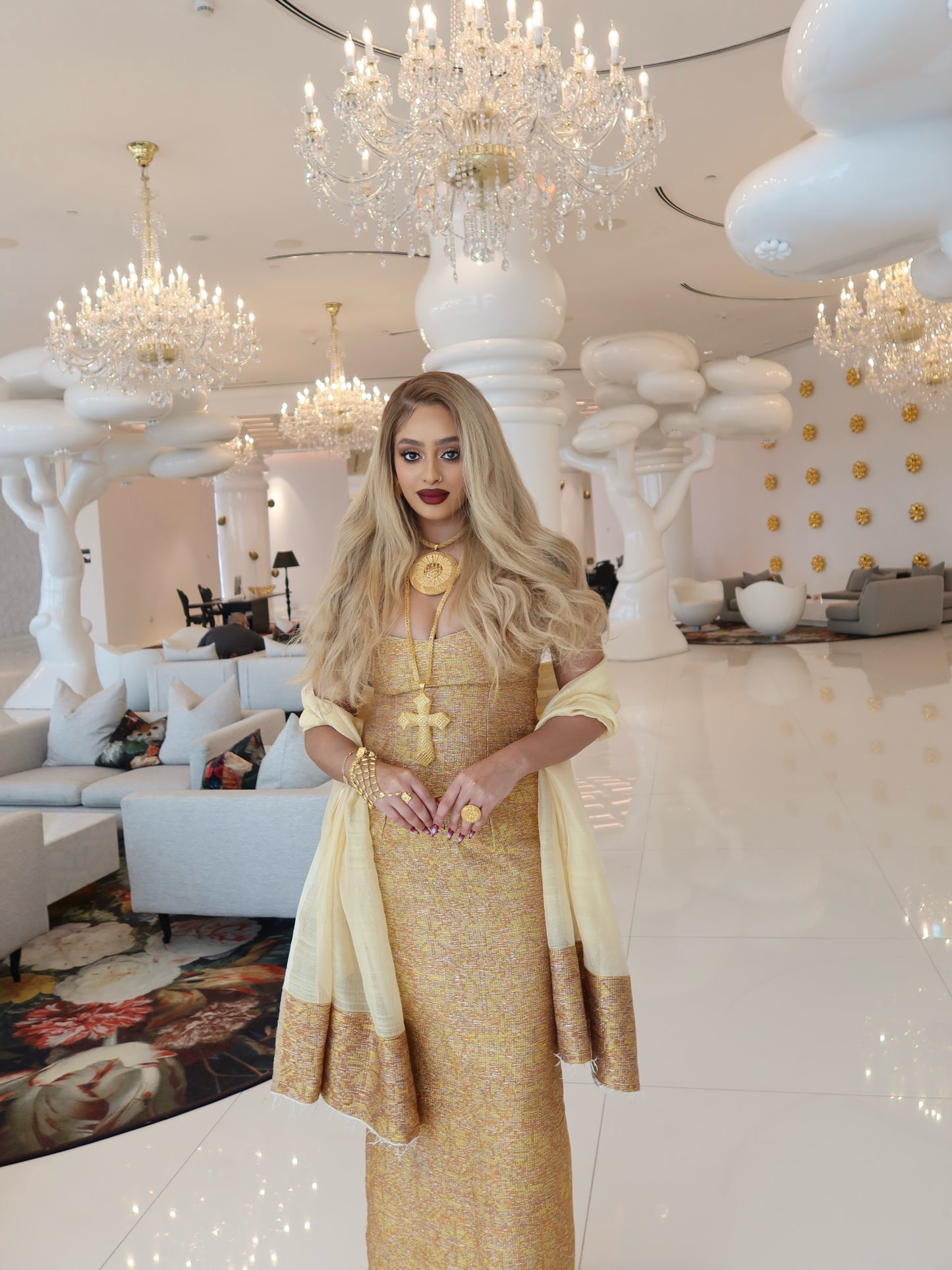 A woman stands in a luxurious, high-ceilinged lobby wearing a shimmering gold and cream traditional Habesha kemis with draped sleeves. The background features elegant white cloud-like pillars, multiple crystal chandeliers, and modern white seating.
