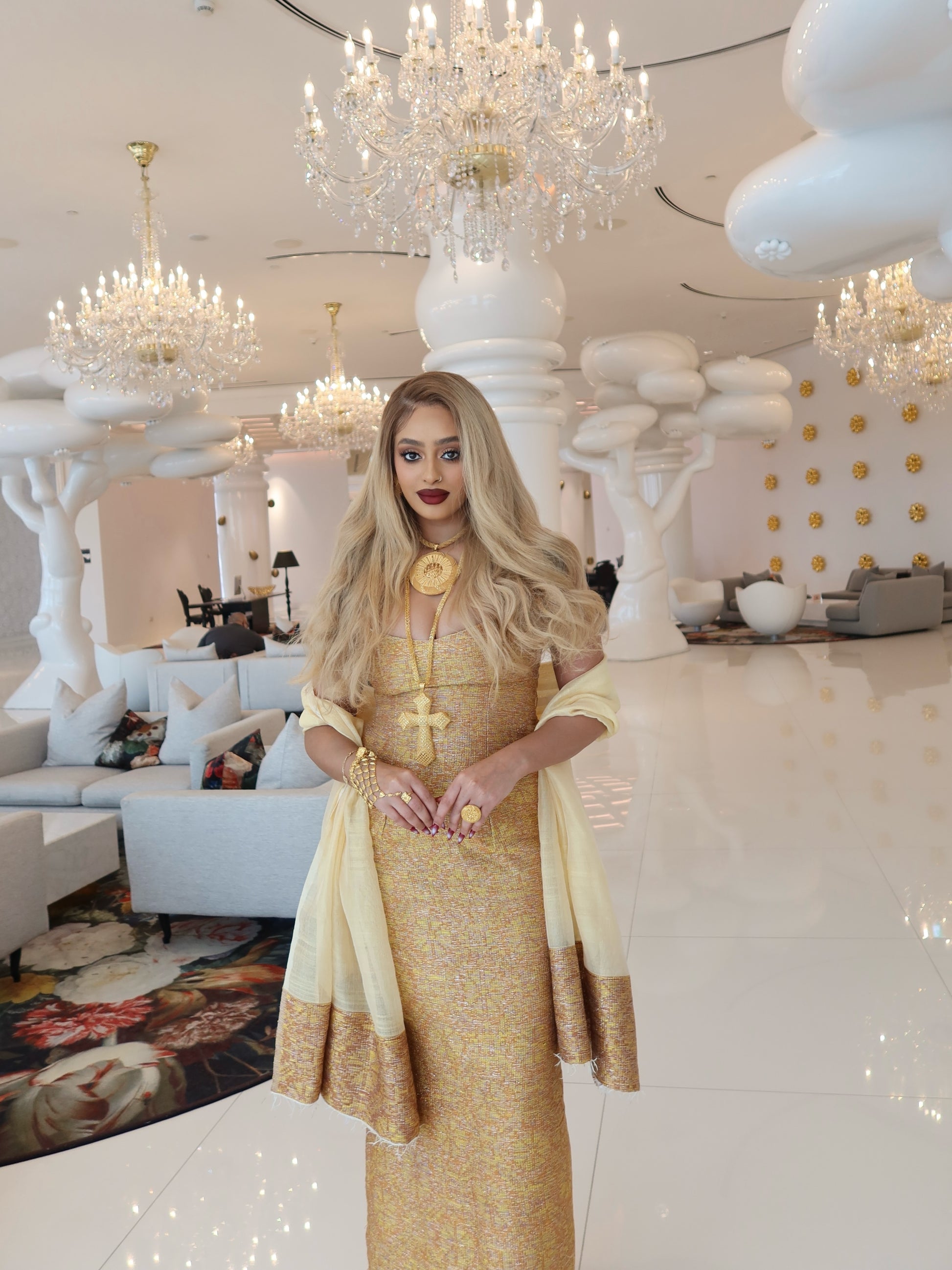 A woman stands in a luxurious, high-ceilinged lobby wearing a shimmering gold and cream traditional Habesha kemis with draped sleeves. The background features elegant white cloud-like pillars, multiple crystal chandeliers, and modern white seating.