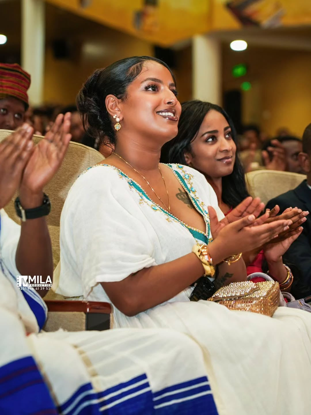 Woman in traditional attire clapping at an event with 'MILA' branding.