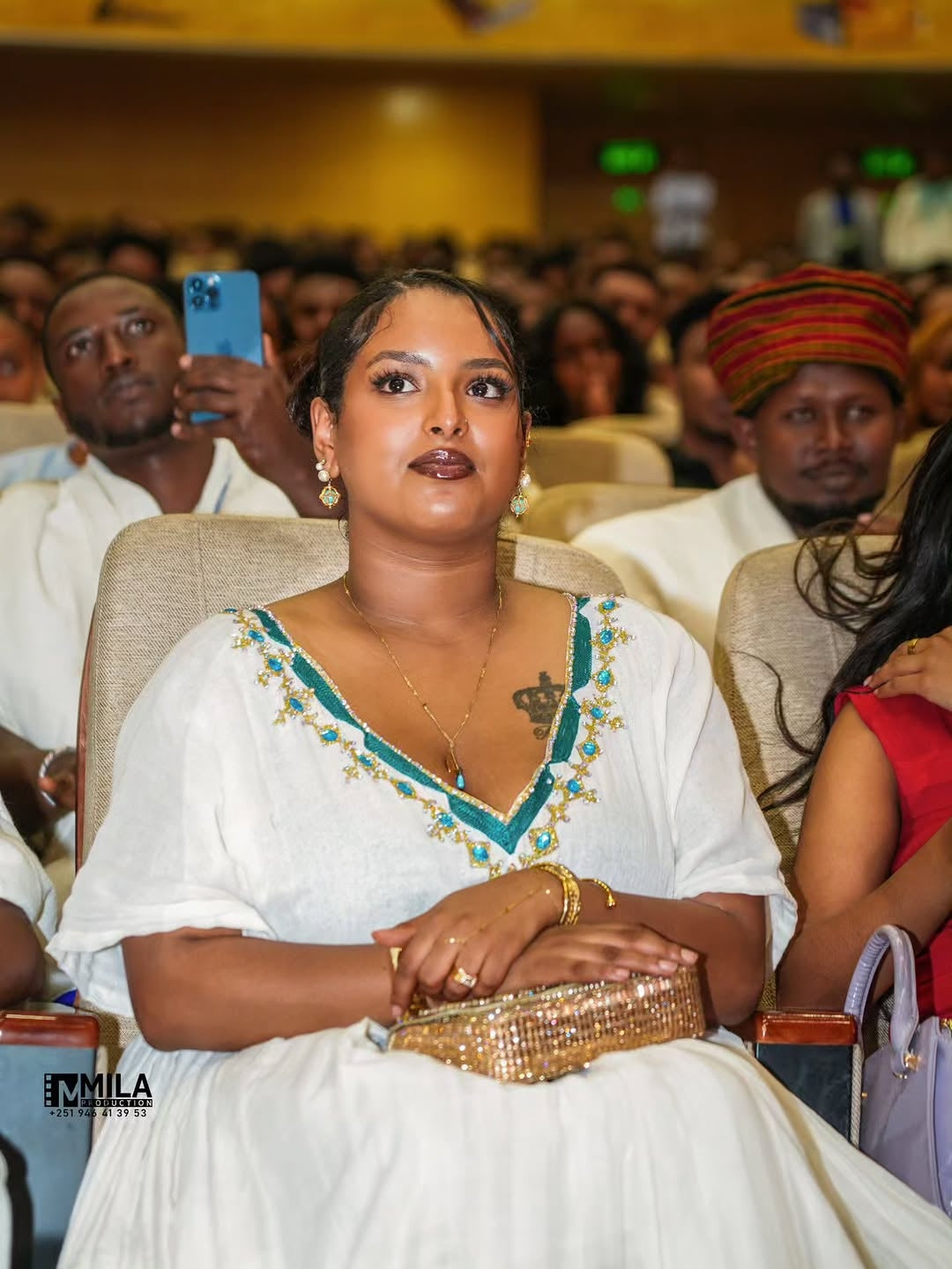 Woman in a white dress with green details sitting in an audience at an event.