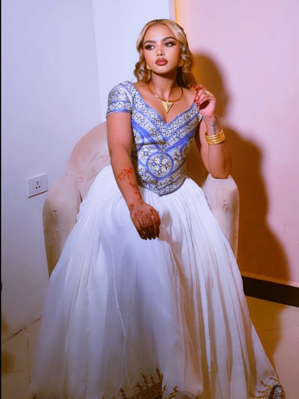 A woman seated in a pink armchair wearing an embellished blue and silver Ethiopian clothes with a voluminous white skirt, photographed indoors against a pale wall.