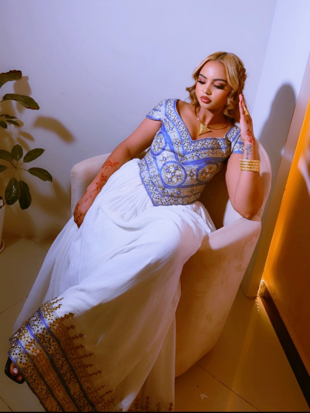 A woman reclining in a beige armchair wearing a traditional Ethiopian clothes outfit with a blue and gold embroidered blouse and a white skirt with gold border detailing.