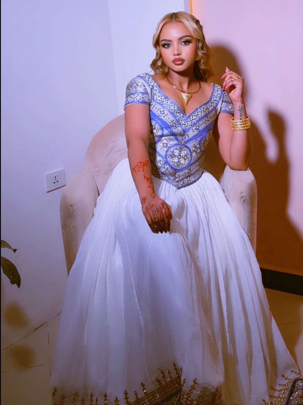 A woman seated in a white armchair wearing a traditional bridal Ethiopian clothes with a heavily embroidered blue and silver bodice and a voluminous white tulle skirt. She is accessorized with gold jewelry, including a necklace and bangles.