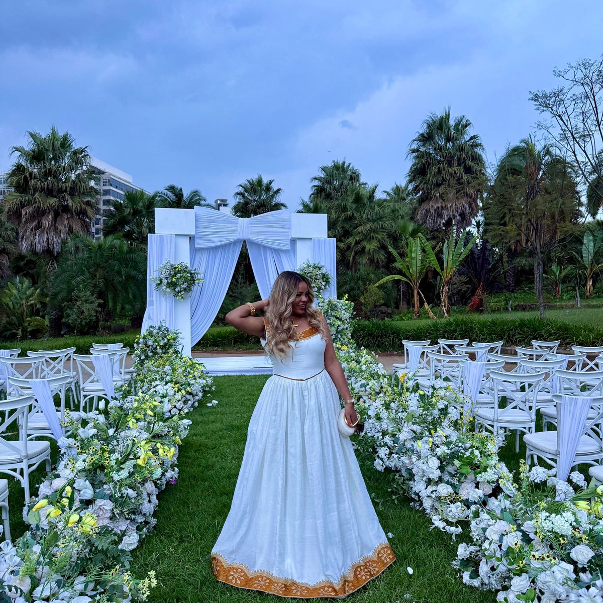 A woman stands on a grassy outdoor aisle wearing a white sleeveless maxi Ethiopian national dresses with gold trim, posing in front of a white draped wedding arch surrounded by white chairs and floral arrangements.